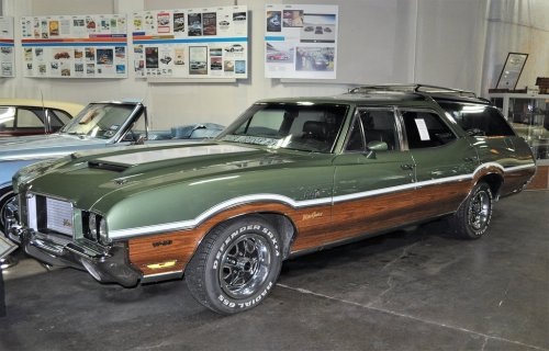 A vintage green station wagon with wood paneling, displayed in a car museum with various posters in the background.