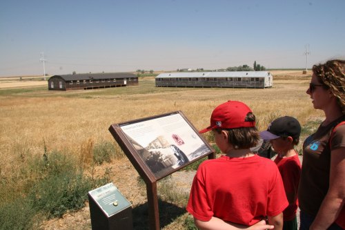 A woman and two children observe a historical sign in a field, with old buildings in the background under a clear sky.