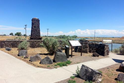 A stone monument with informational signs near a river, surrounded by sparse vegetation and a clear blue sky.
