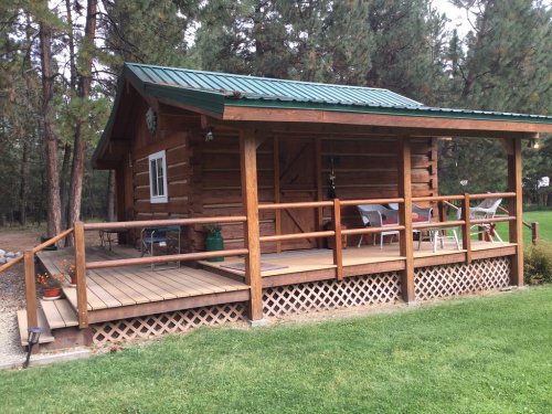 A cozy log cabin with a green metal roof, featuring a wooden porch and chairs, surrounded by trees and grass.