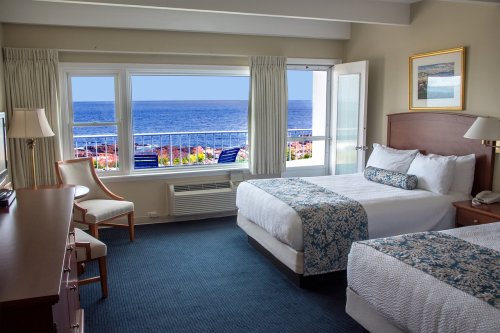 Bright hotel room with two beds, a chair, and a stunning ocean view through large windows.
