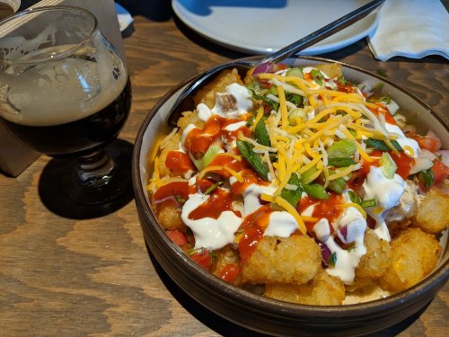 A bowl of loaded tater tots topped with cheese, sour cream, salsa, and green onions, alongside a glass of dark beer.