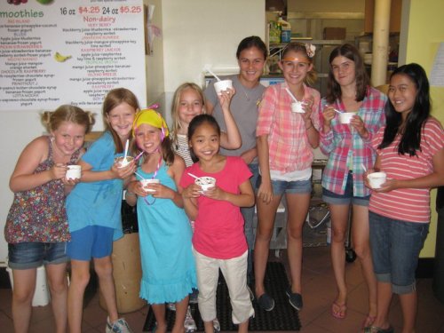 A group of nine girls of varying ages smiles while holding cups of ice cream in a colorful shop.