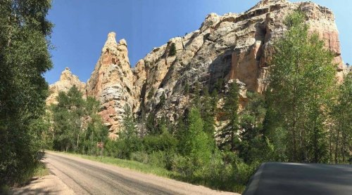 Panoramic view of towering rock formations beside a gravel road, surrounded by lush greenery under a clear blue sky.