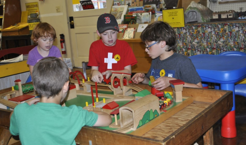 Four children play with a wooden train set on a table, focused on building tracks and structures.