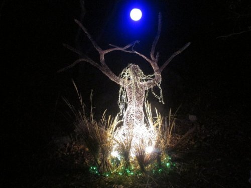 A glowing, abstract tree sculpture with lights, set against a dark background and a bright blue moon above.