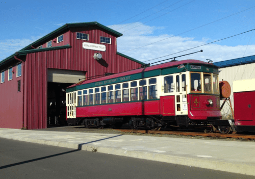 A vintage red and cream trolley parked outside a red barn-like building on a sunny day.