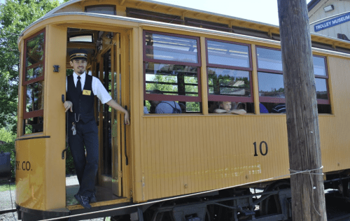 A conductor stands at the entrance of a vintage yellow trolley, with passengers visible inside.