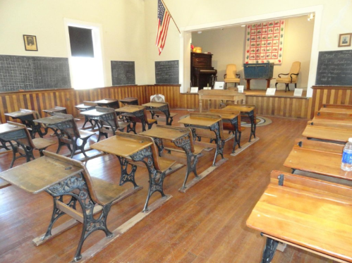 Historic classroom with wooden desks, chalkboards, and an American flag, featuring a stage at the front.