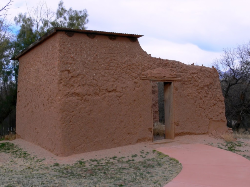A small adobe structure with a flat roof, featuring a wooden door and surrounded by sparse vegetation.