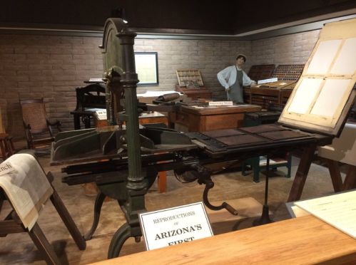 A historic printing press displayed in a museum, with wooden furniture and a mannequin in the background.