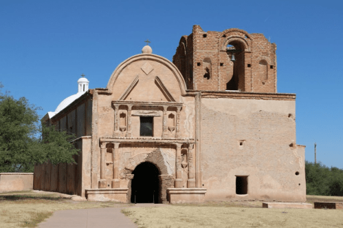 Historic adobe church with a bell tower, set against a clear blue sky and surrounded by sparse vegetation.