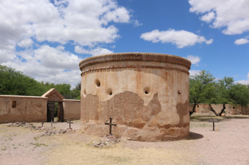 Historic round adobe structure with a textured exterior, surrounded by a desert landscape and blue sky with clouds.