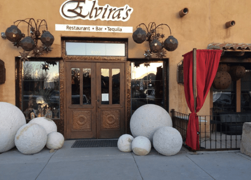 Entrance of Elvira's Restaurant with wooden doors, hanging lanterns, and large round stones at the base. Red curtain to the side.