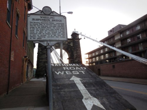 Wheeling Suspension Bridge sign with a view of the bridge and road leading west, surrounded by historic buildings.