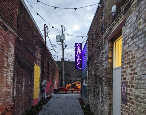 Narrow alleyway with brick walls, string lights, and a neon sign reading "Unplugged" at the end.