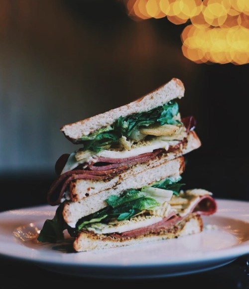 A tall sandwich stacked with layers of lettuce, meats, and bread, served on a white plate with a blurred background.