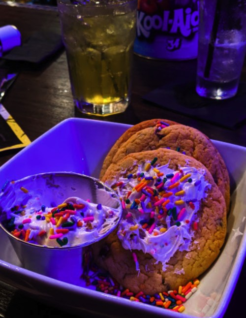 A bowl with two large cookies topped with frosting and colorful sprinkles, alongside a scoop of frosting. Drinks in the background.