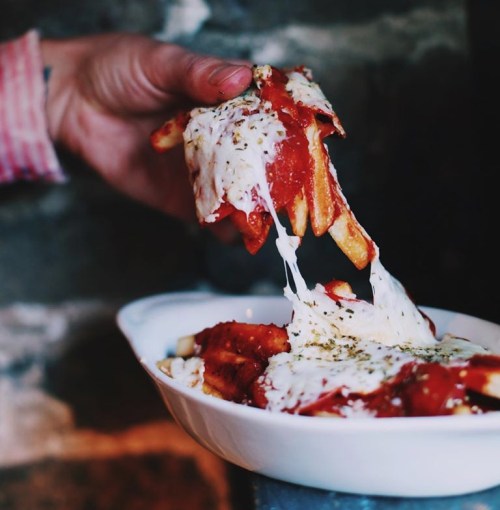A hand lifts cheesy fries topped with marinara sauce from a white dish, with herbs sprinkled on top.
