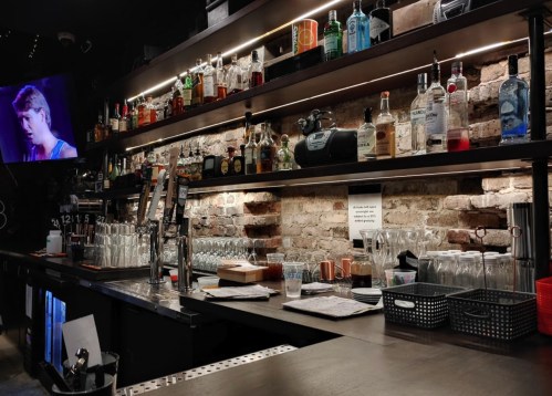 A bar with shelves of various liquor bottles, glasses, and a TV screen in a dimly lit setting.