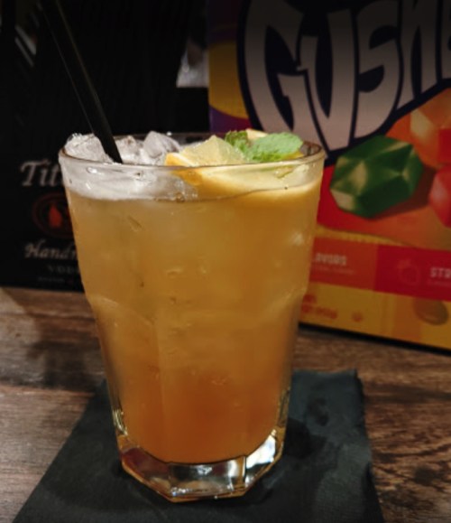 A refreshing cocktail with ice, lemon slices, and mint, served in a glass on a black napkin, with colorful candy in the background.
