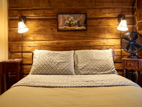 Cozy bedroom with a wooden wall, a neatly made bed, bedside lamps, and a fan. A floral painting hangs above the bed.