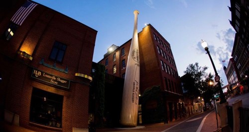 A large baseball bat sculpture stands outside the Louisville Slugger Store at dusk, with buildings and streetlights nearby.