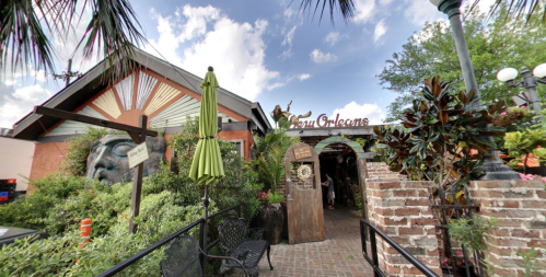 Entrance to a vibrant restaurant with tropical plants, a large sculpture, and a welcoming sign under a blue sky.