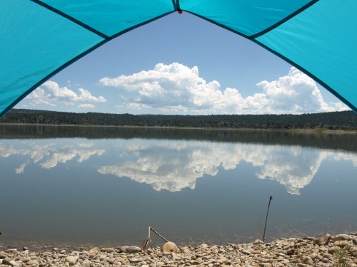 View of a calm lake reflecting clouds, framed by a blue tent canopy and rocky shoreline.