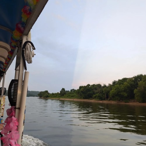 A scenic view from a boat, showing calm waters and lush greenery along the riverbank under a cloudy sky.