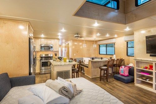 Interior of a modern RV featuring a kitchen, dining area, and living space with a bed and large windows.