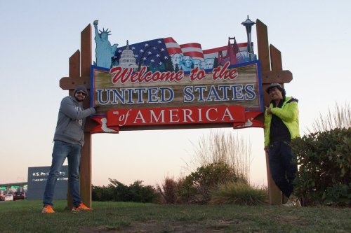 Two people pose beside a colorful "Welcome to the United States of America" sign, with iconic landmarks illustrated.