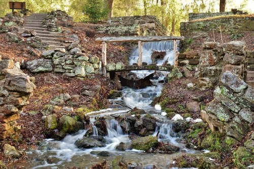 A serene stream flows over rocks, with a small wooden bridge crossing it, surrounded by lush greenery and stone walls.