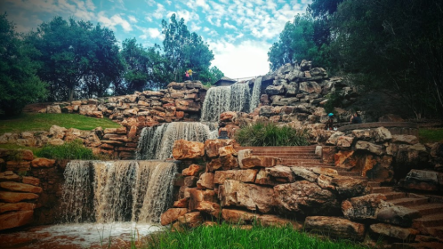 A scenic waterfall cascading over rocks, surrounded by greenery and trees under a partly cloudy sky.