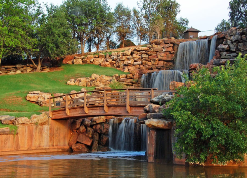 A serene park scene featuring a wooden bridge over a waterfall, surrounded by lush greenery and rocky terrain.