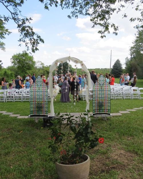 A wedding ceremony setup with guests gathered under a floral arch, surrounded by greenery and white chairs.
