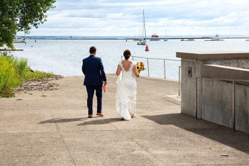 A couple walks hand in hand by the water, with a bouquet and boats in the background on a cloudy day.