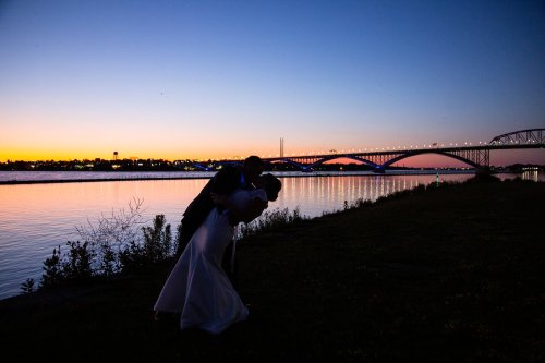 A couple embraces at sunset by a river, with a bridge silhouetted against a colorful sky.