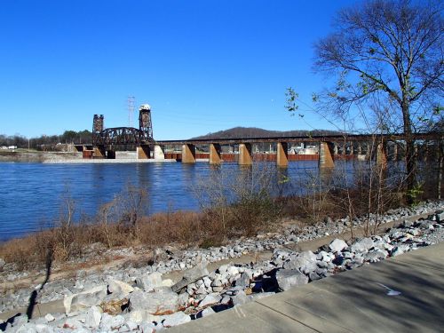 A river scene featuring a bridge with towers, rocky shoreline, and clear blue sky in the background.