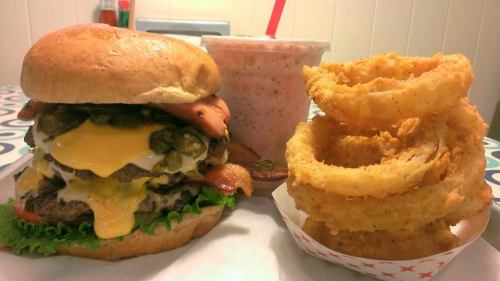 A large burger with cheese, bacon, and toppings, served with onion rings and a milkshake on a colorful table.