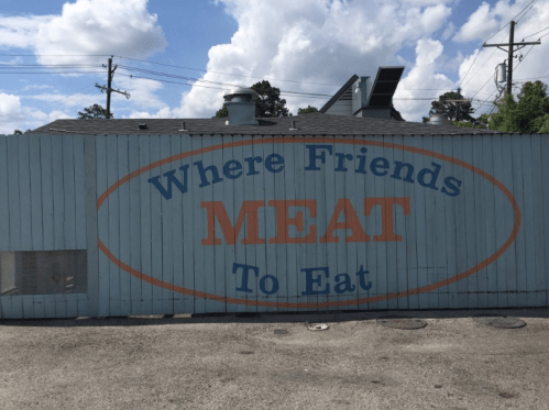 A wooden fence with a sign that reads "Where Friends MEAT To Eat" against a blue sky with clouds.