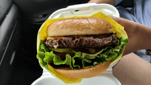 A hand holding a juicy hamburger with lettuce, tomato, pickles, and cheese, wrapped in yellow paper.