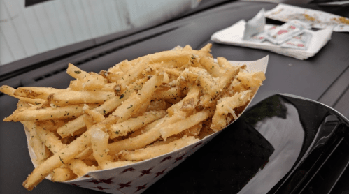 A serving of golden, crispy French fries in a paper tray, sprinkled with herbs, on a car dashboard.