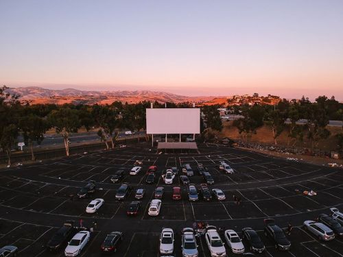 Aerial view of a nearly empty drive-in movie theater at sunset, with cars parked in front of a large blank screen.