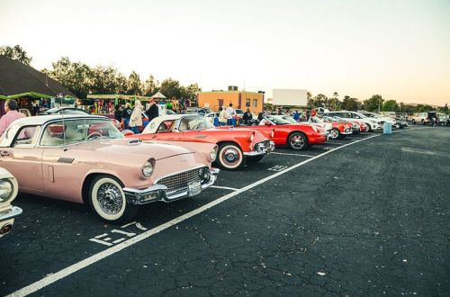 A row of classic cars in a parking lot during sunset, with people mingling nearby.