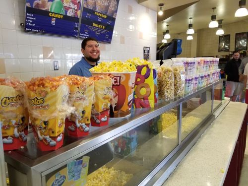 A smiling employee stands behind a counter filled with colorful popcorn and snacks at a movie theater concession stand.