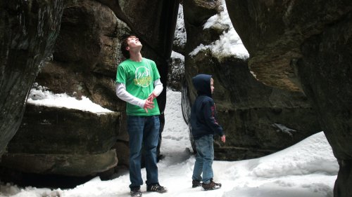 Two boys in winter clothing explore a snowy rocky area, looking up at the towering rock formations above them.