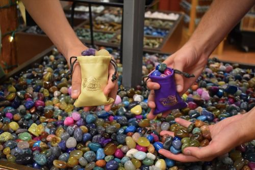 Two hands hold colorful gemstone bags above a pile of various polished stones in a shop.