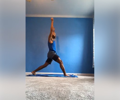 A person in a blue tank top and shorts performs a yoga pose on a mat in a room with blue walls and carpet.
