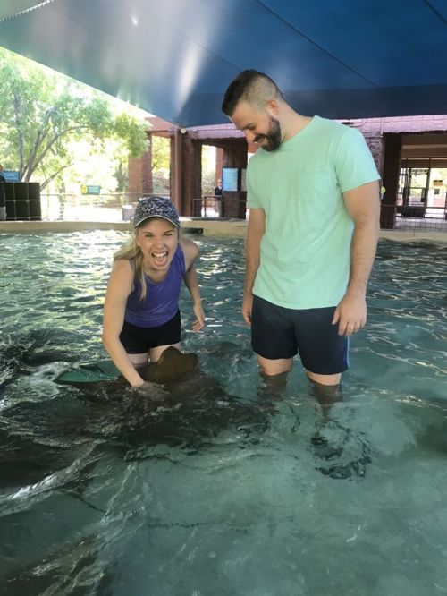A woman and a man stand in shallow water, smiling as they interact with a stingray.